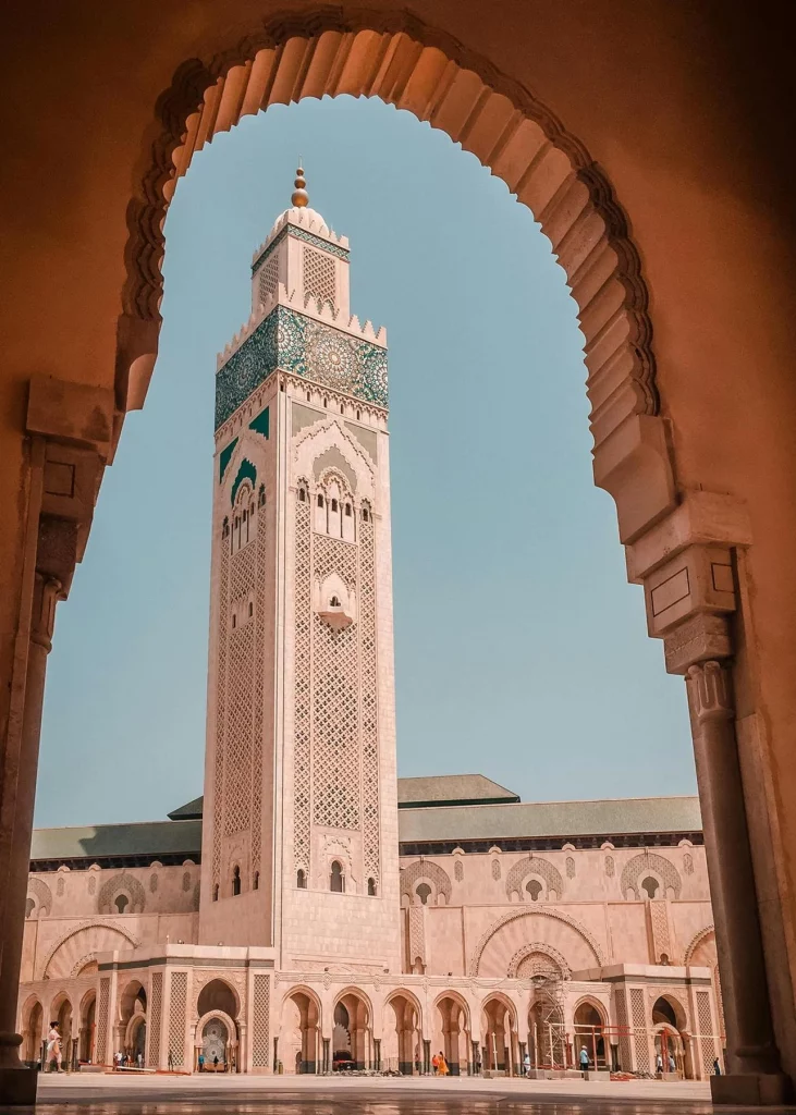A low-angle shot of the towering minaret of the Hassan II Mosque in Casablanca, framed perfectly by a large, ornate stone archway in the foreground. The massive cream-colored minaret features intricate geometric carvings and a prominent band of green and blue mosaic tilework near its top. The main mosque building extends out below with a long arcade of arches under a pale, clear blue sky.