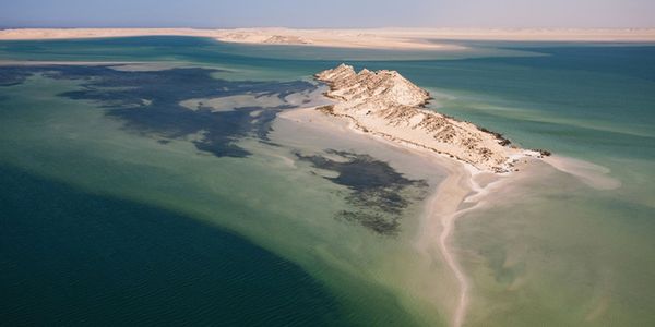 High-angle aerial view of Dragon Island (Île du Dragon) in the Dakhla lagoon, showing a stark, rocky sand island surrounded by shallow, shifting turquoise and deep green waters under a clear sky.